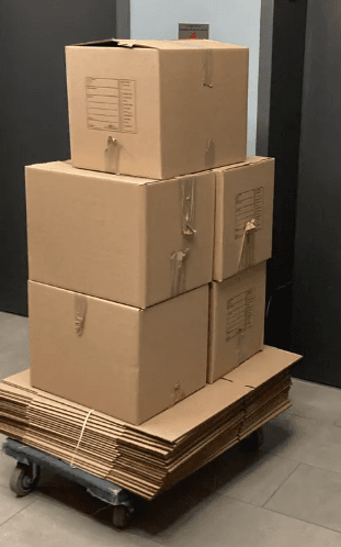 Five cartons stacked on a flat dolly, riding on top of a bundle of flattened boxes, in a corridor by the lift