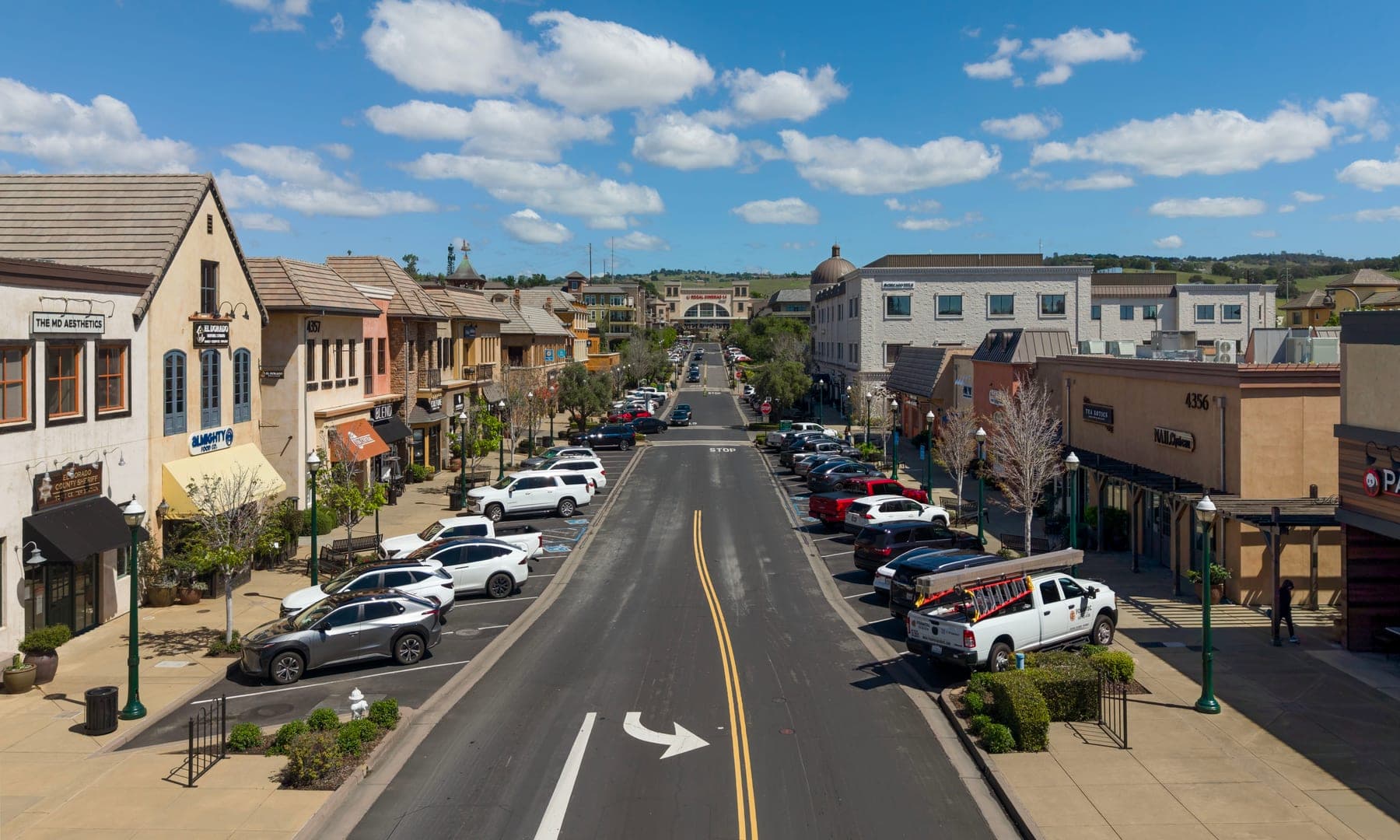 The main street of El Dorado Hills Town Center — a wide two-lane road, angle parking on both sides, tile-roofed shopfronts and the dry foothills behind