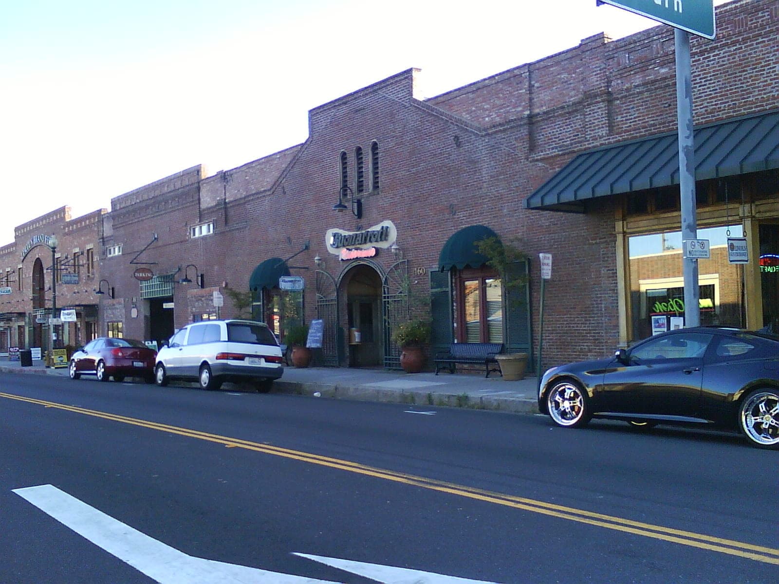 A block of downtown Lincoln in evening light — single-storey red-brick storefronts under green awnings, cars at the kerb and a wide asphalt road across the foreground